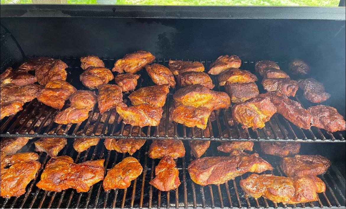 482029591_1097167909091239_6493213131961913580_n Smoked chicken pieces cooking on a grill, showcasing a variety of textures and rich, dark seasoning. The image captures multiple layers of chicken on the grill grates, emitting smoke, indicating a flavorful barbecue process. Perfect for food enthusiasts and grilling aficionados.