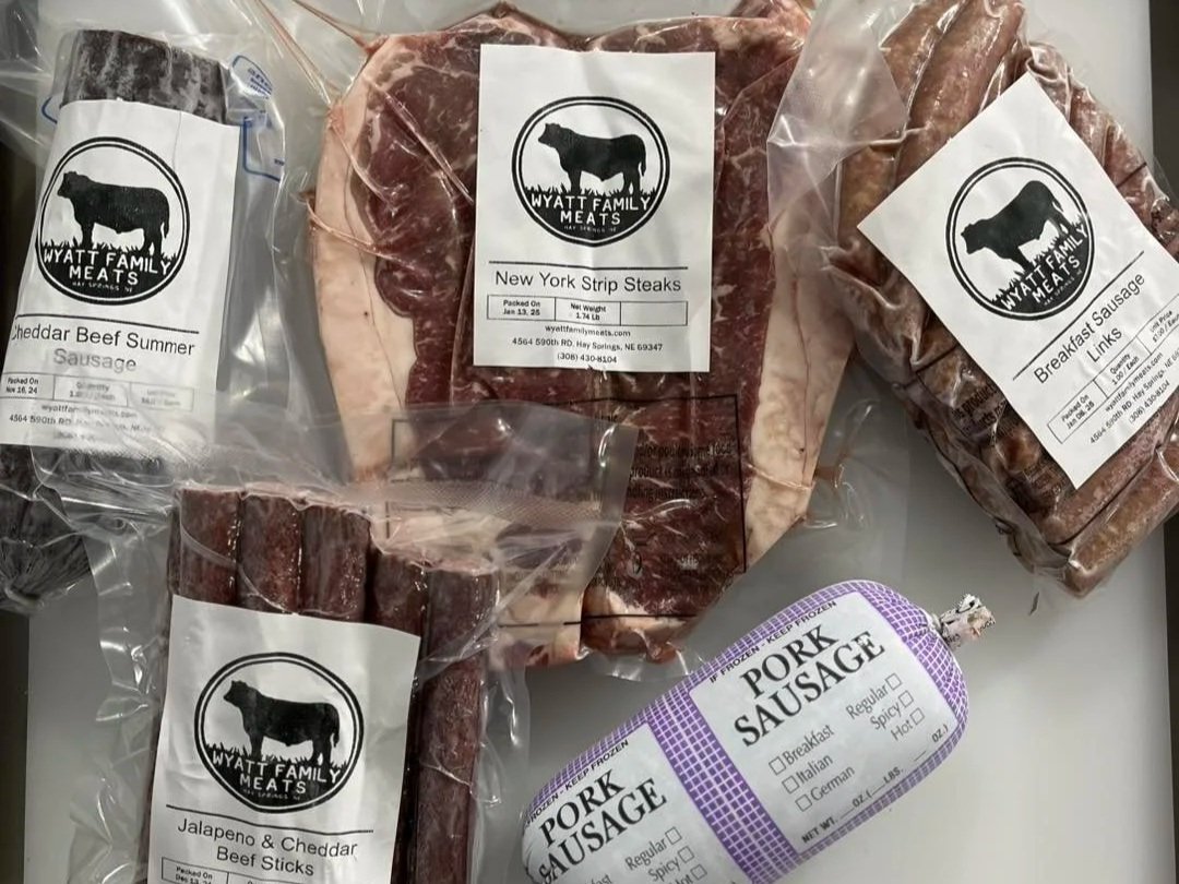 Assorted meat products from Wyatt Family Meats, featuring New York strip steaks, cheddar beef summer sausage, jalapeno and cheddar beef sticks, and breakfast sausage links, all packaged and labeled for freshness.
