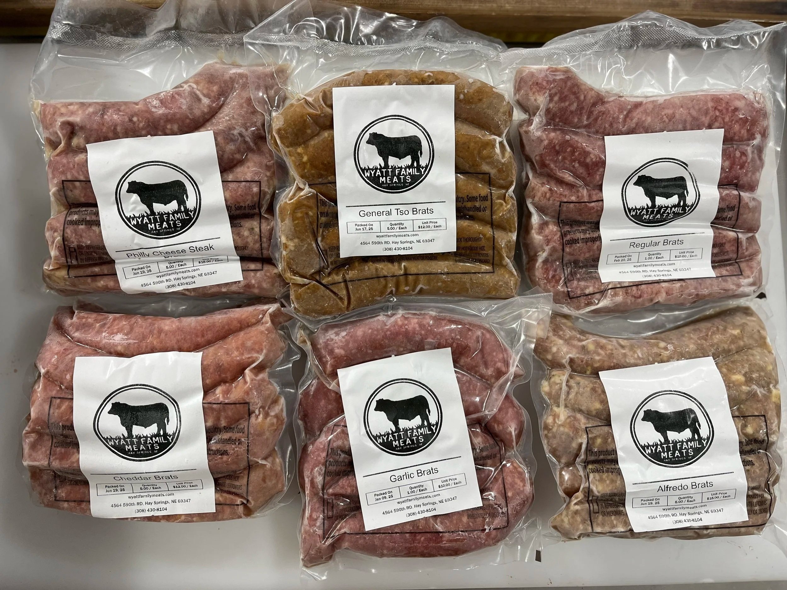 Amanda+Newman-Wyatt+Family+Meats+-+IMG_7917 Variety of packaged sausages from Wyatt Family Meats, including Philly Cheese Steak, General Tso Brats, Regular Brats, Cheddar Brats, Garlic Brats, and Alfredo Brats, displayed on a white surface. Each package features a clear label with product information and branding.