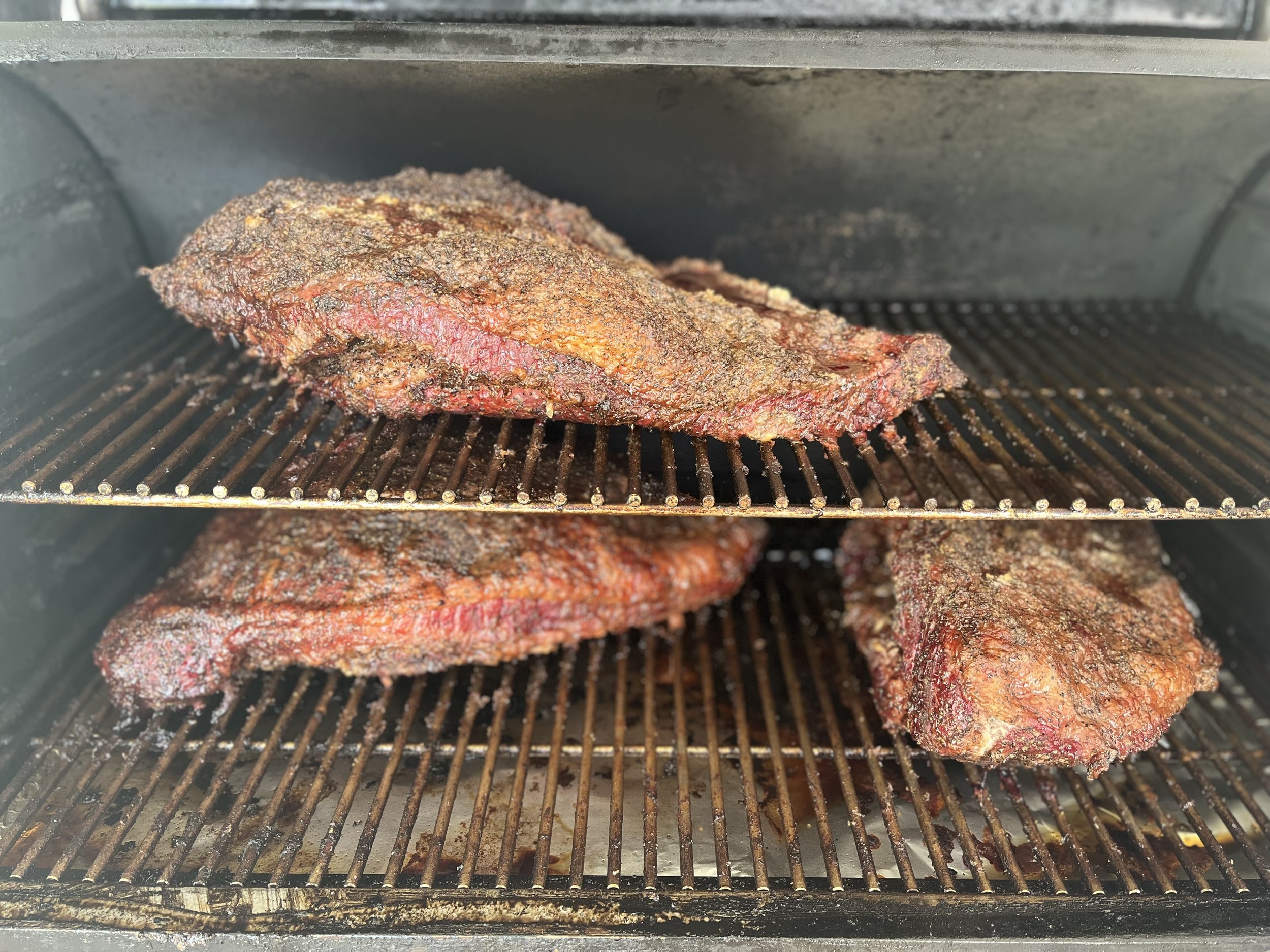 Brisket+on+Smoker Smoked brisket cooking in a barbecue smoker, showcasing three large cuts with a dark, flavorful crust. The meat is positioned on metal racks, with juices visible on the grill surface, highlighting the slow-cooked barbecue technique.