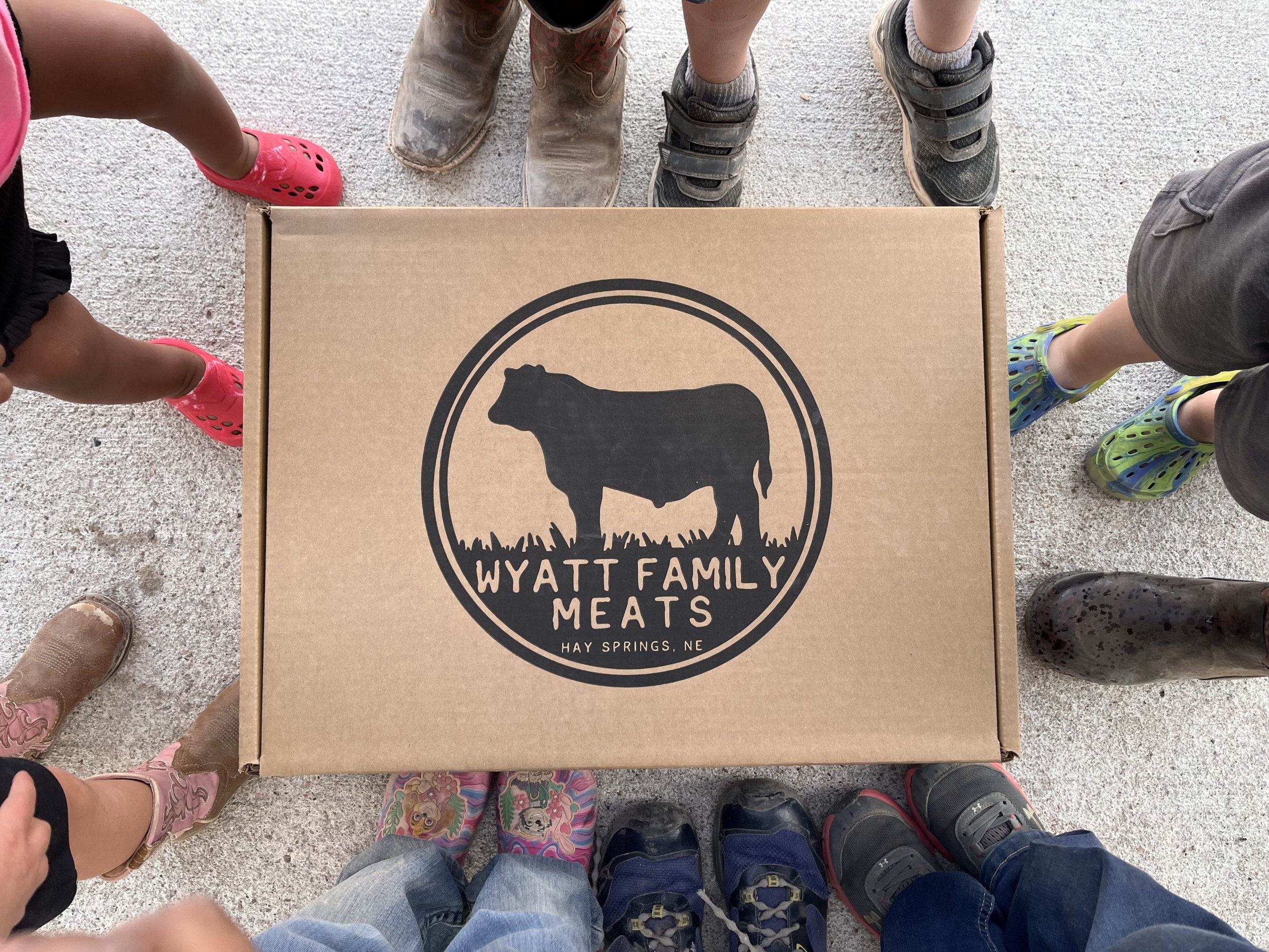 Group of children and adults standing around a cardboard box labeled "Wyatt Family Meats" from Hay Springs, NC, showcasing local meat products. The image highlights the diverse footwear of the group, emphasizing a rural, family-oriented atmosphere.