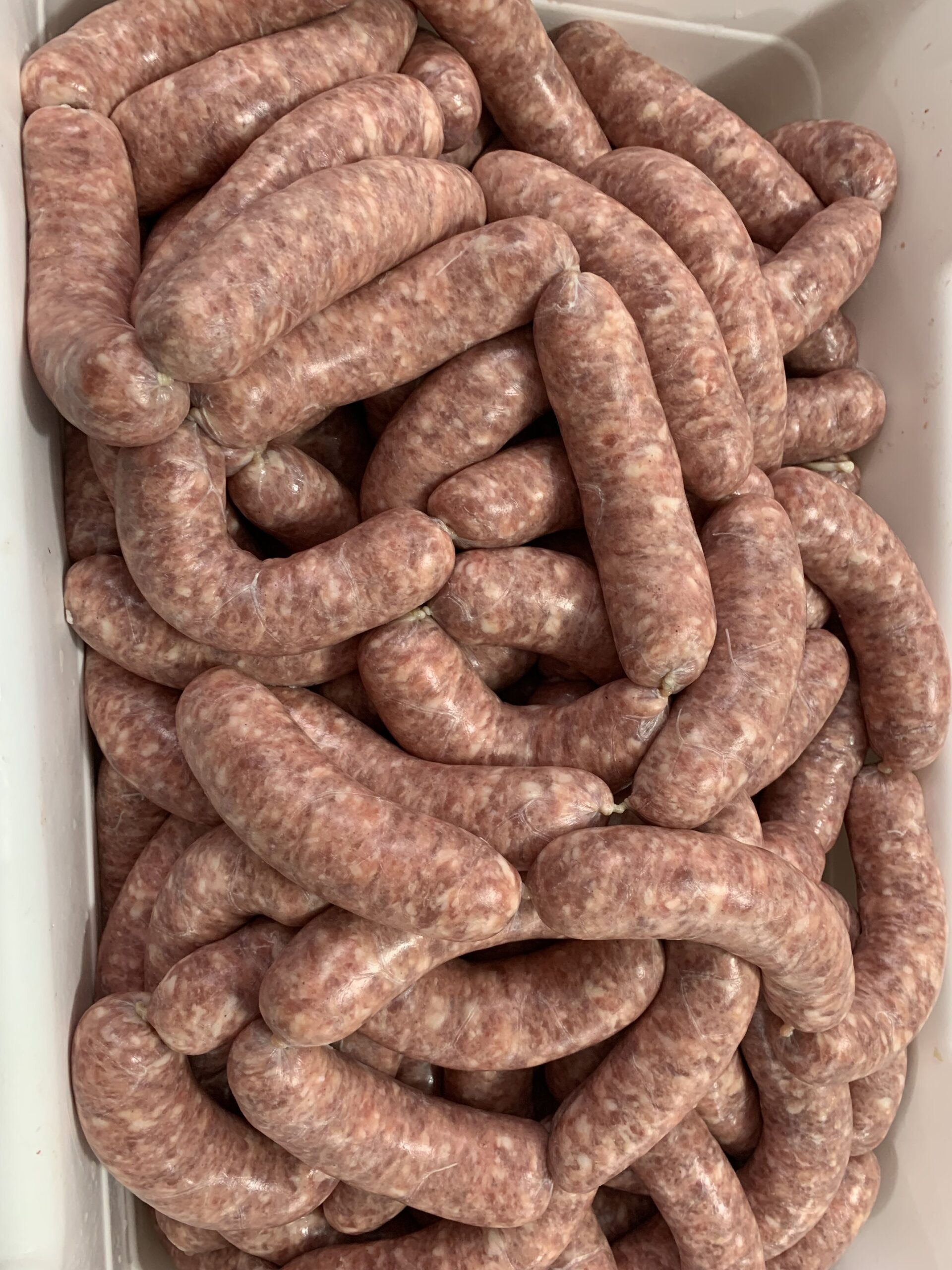 lug+of+brats Freshly made sausages piled together in a white container, showcasing their natural pink color and casing. Ideal for grilling or cooking, these sausages are a popular choice for barbecues and meals.
